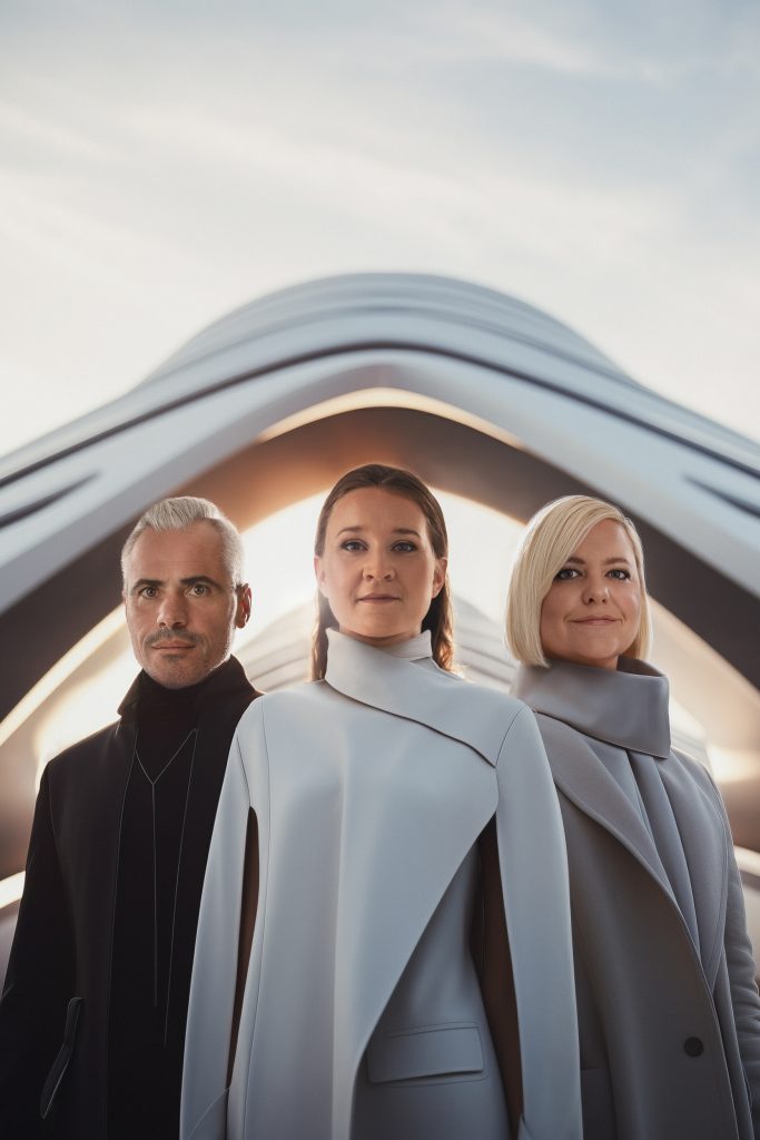 Zwei Frauen und ein mann schauen direkt in die Kamera, alle drei haben einen Mantel an, sie schauen alle drei in die Kamera und alles drei schauen fröhlich aus