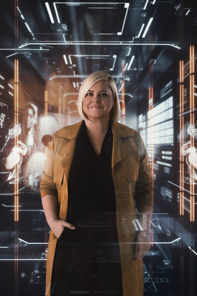 Frau steht in einem engen Gang und rum um die Frau herum kann man lauter elektronische Sachen erkennen, die Frau schaut in die Kamera und lächelt, sie hat auch ihre rechte Hand in die Hosentasche