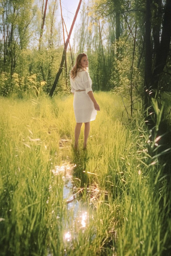 Eine Frau steht in einem Wald wo sehr viel Gras und Wasser am Boden sind, sie geht vorwärts dreht sich aber Richtung Kamera um und schaut direkt in die Kamera