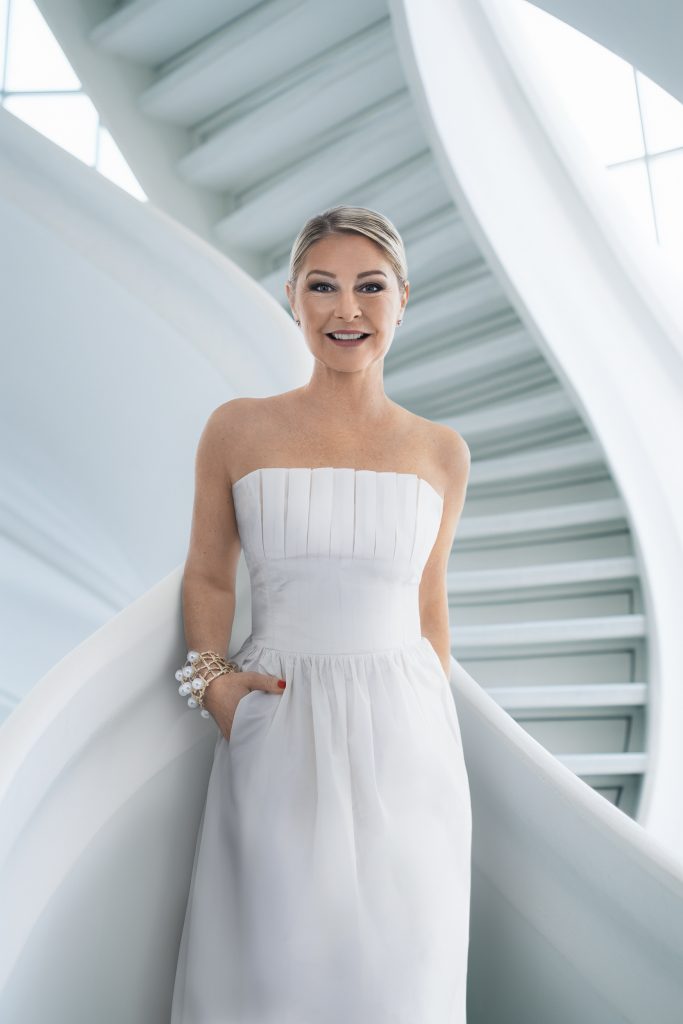 Eine Frau steht vor einer riesigen Treppe, sie schaut direkt in die Kamera und lächelt, sie hat eine Hand in ihrem Kleid eingesteckt