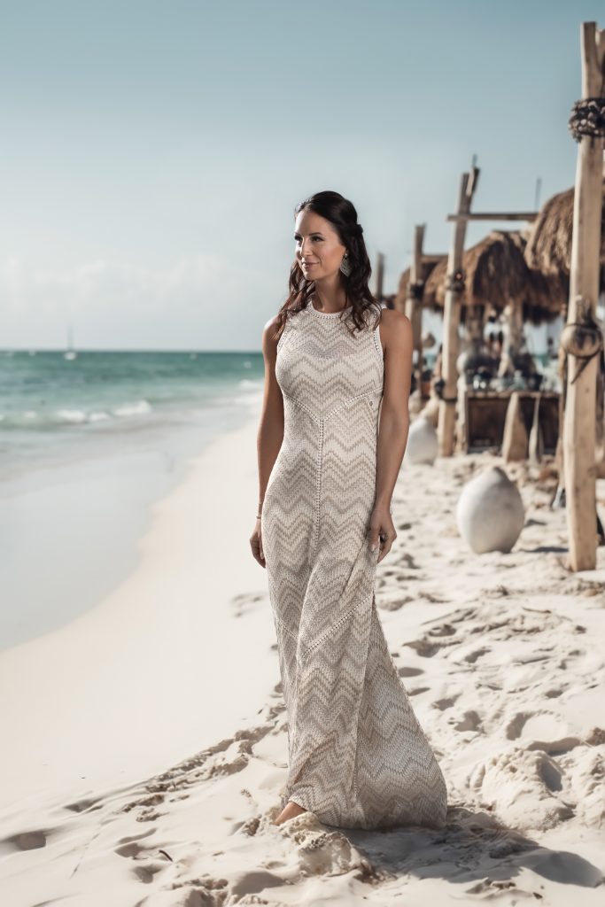 Eine Frau geht auf dem Sand neben dem Meer, sie trägt ein langes weißes Kleid und schaut an der Kamera vorbei, sie lächelt Richtung Sonne