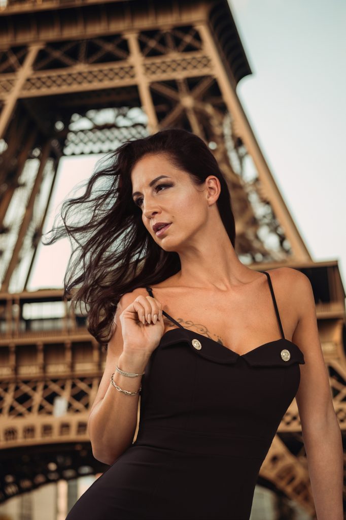 Frau steht vorm Eiffelturm, sie schaut an der Kamera vorbei, sie trägt ein Kleid und hat ein Tatto im Dekoltee Bereich