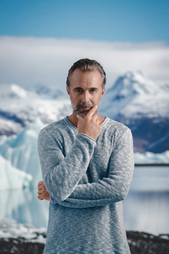 Mann steht vor ein paar Eisbergen, er schaut direkt in die Kamera mit einem fragenden Gesichtsausdruck, er stützt seine rechte Hand im Gesicht ab
