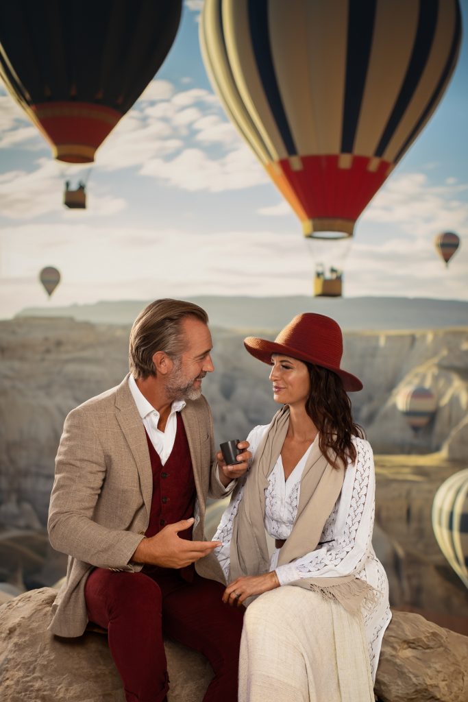 Eine Frau und ein Mann sitzten auf einem Felsen und trinken Kaffee, im Hintergrund fliegen sechs Heißluftballons über einigen Felsen