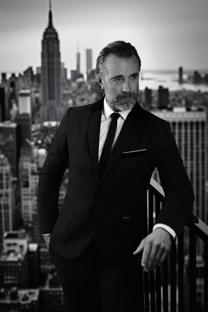 schwarz-weiß Bild, Mann steht auf einem Hochhaus und lehnt sich an ein Geländer, er schaut an der Kamera vorbei und im Hintergrund sieht man ganz viele Großgebäude