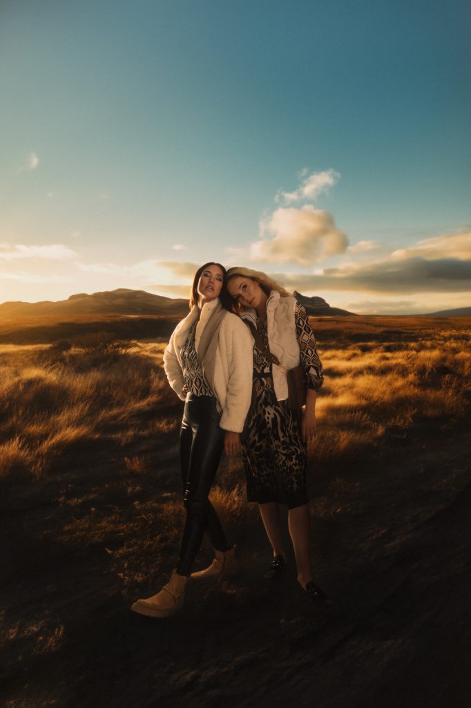 Zwei Frauen stehen auf einem Weg, links und rechts ist Wiese, eine der beiden Frauen schaut in die Kamera und die andere hat ihre Augen geschlossen