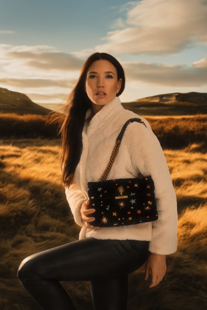 Frau steht in einem Feld und schaut in die Kamera, sie hat eine Handtasche umhängen und hält diese in der Hand