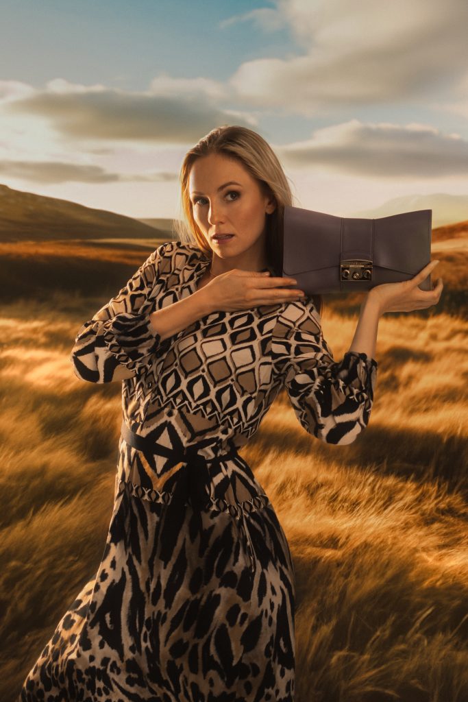 Frau steht in einem Feld, sie hält sich eine Tasche direkt neben den Hals, sie schaut an der Kamera vorbei