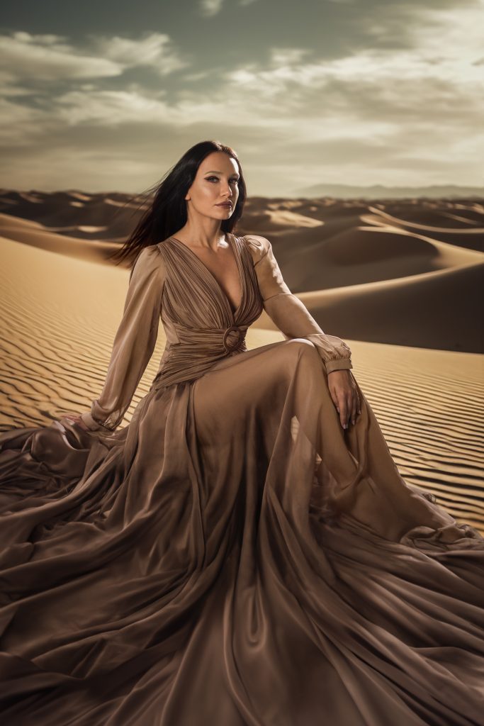 Frau sitzt in der Wüste auf einem sandhügel, sie schaut emotionslos in die Kamera, sie trägt ein langes Kleid und stützt sich mit der rechten Hand ab