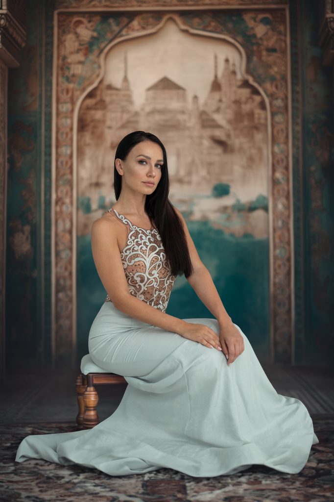 Frau sitzt auf einem Hocker, im Hintergrund ist ein Wand Mandala, sie trägt ein türkises Kleid und schaut in die Kamera